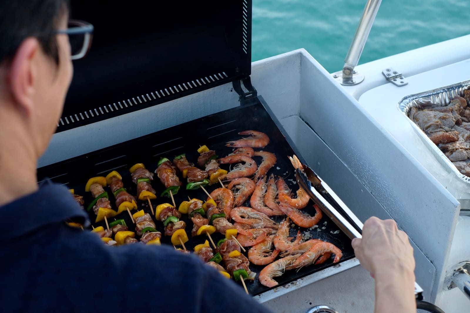 A guest turning prawns and chicken skewers with tongs on an open gas grill at the side deck of a charter boat, a foil tray of marinated meat resting beside the grill and green water beyond the rail