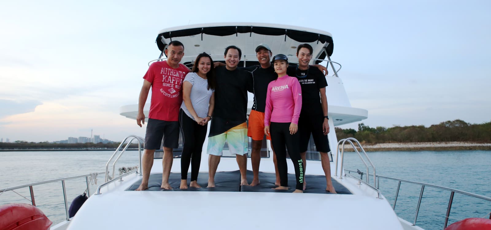 The founders of B.O.B Marine with members of their crew, standing arm in arm on the bow of a moored yacht at dusk, with the Singapore skyline low on the horizon behind them