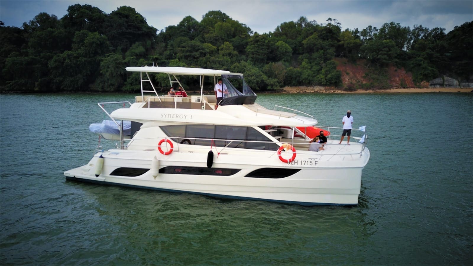 Synergy 1 at anchor off a wooded Singapore island, guests on the foredeck and the flybridge, with her name on the hull side